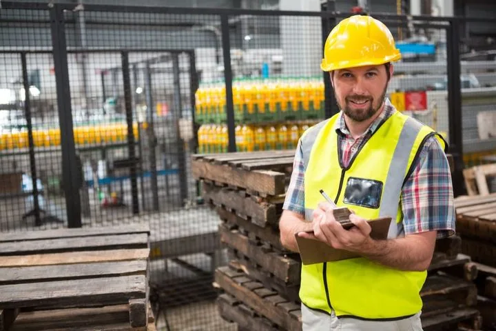 Mitarbeiter mit Schutzhelm und Warnweste prüft im Lager Arbeitsplätze anhand einer Checkliste