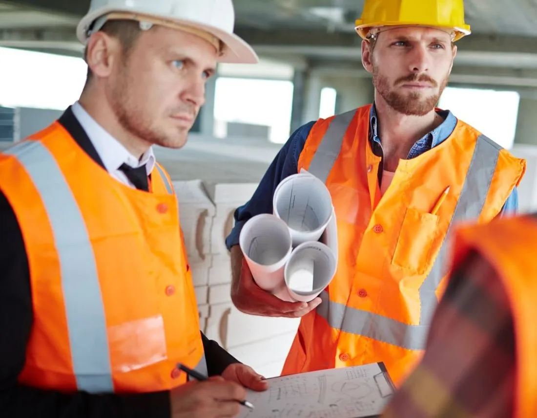 Zwei Mitarbeiter mit Helm und Warnweste besprechen Pläne in einer Halle